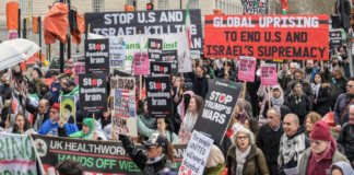 Protesters wave flags and banners reading “Stop Bombing Iran” and “Stop Bombing the Middle East” as they gather at Victoria Tower Gardens in London