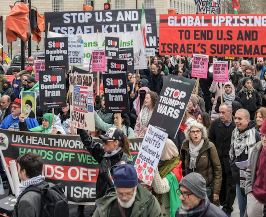 Protesters wave flags and banners reading “Stop Bombing Iran” and “Stop Bombing the Middle East” as they gather at Victoria Tower Gardens in London